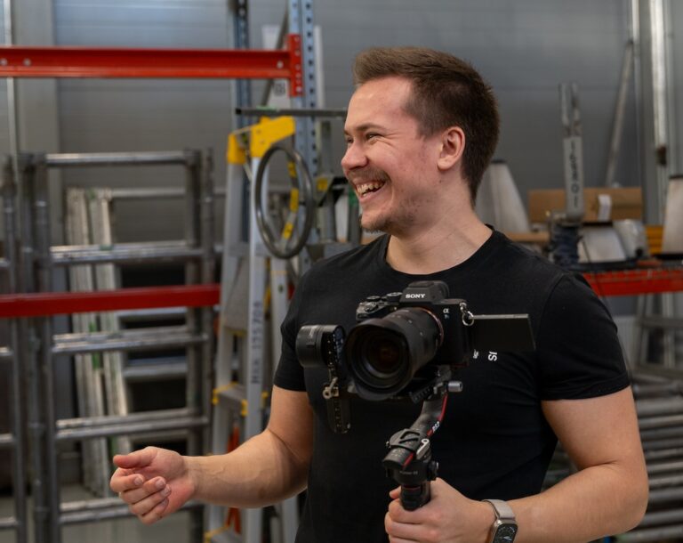 Videokuvaaja William gimbal kädessä hymyilemässä asiakkaan varastotiloissa kuvauksissa.